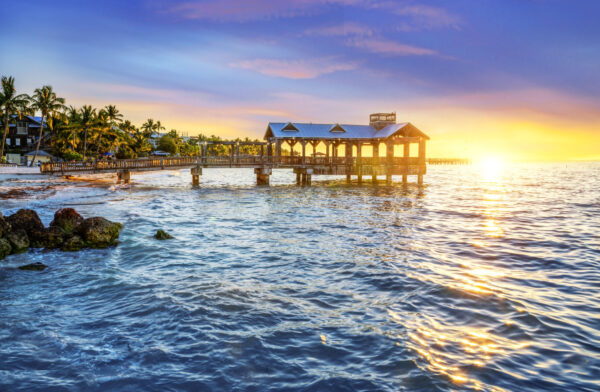 Wooden pier extends into calm ocean at sunset, palm trees lining the shore on the left.